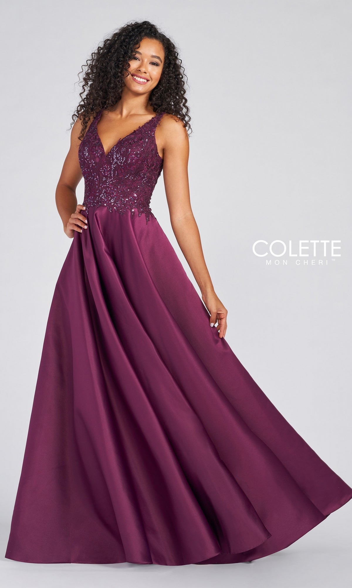 A woman stands against a plain light background wearing a sleeveless, floor-length burgundy V-Neck Satin Prom Dress by Colette by Daphne, featuring an embroidered bodice and flowing skirt.